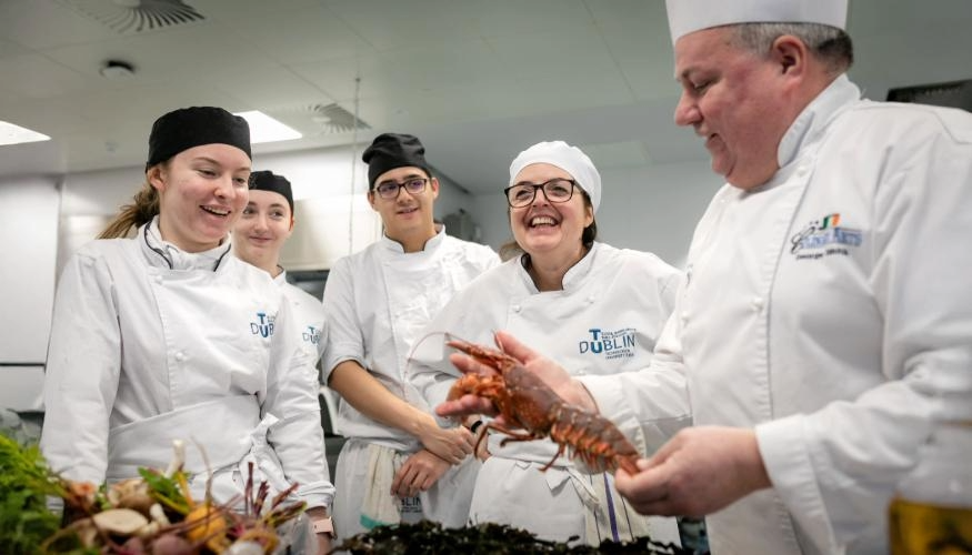 group of students in training kitchen watching chef holding lobster