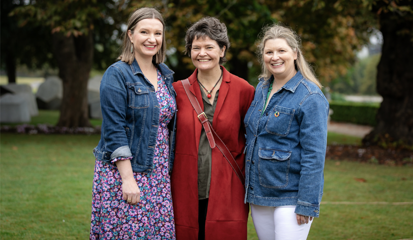 three smiling woman posing together for a photo on a lawn with trees in the background