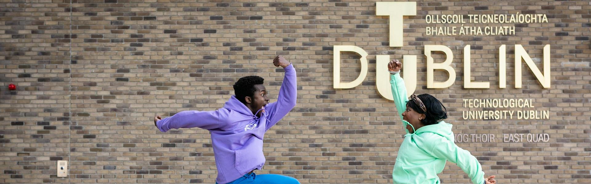 Two students leaping and dancing beside the TU Dublin sign